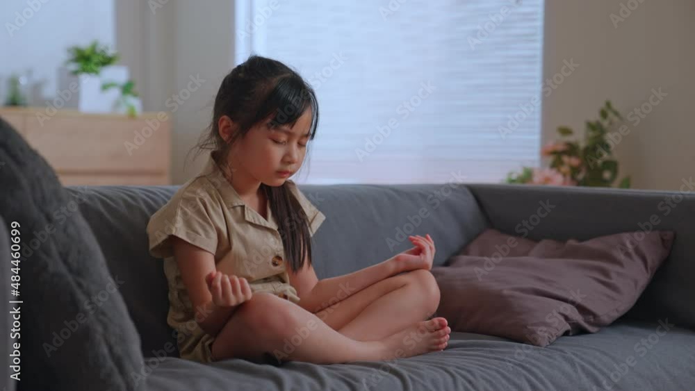 Asian baby girl practicing yoga in morning, siting in lotus pose and meditates on sofa in the room with her eyes closed. Attractive slender girl relaxes plunges into world self discovery. Asian baby girl practicing yoga in morning, siting in lotus pose and meditates on sofa in the room with her eyes closed. Attractive slender girl relaxes plunges into world self discovery.