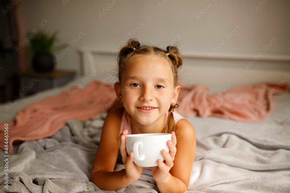 Cute little happy girl with two ponytails in her bed drinking hot tea from white cup Cute little happy girl with two ponytails in her bed drinking hot tea from white cup
