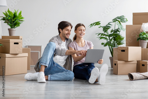 Smiling caucasian young husband and wife look at pc among cardboard boxes with stuff in room