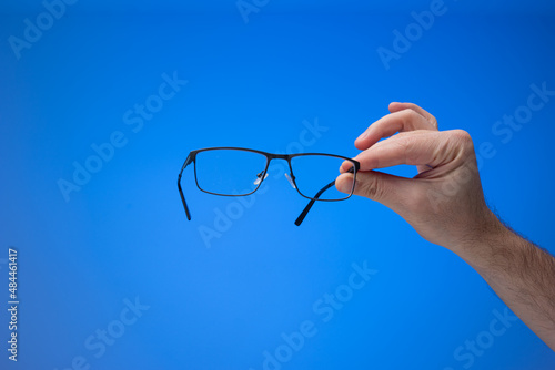 Pair of new plastic framed eyeglasses held in hand by Caucasian male. Close up studio shot, isolated on blue background