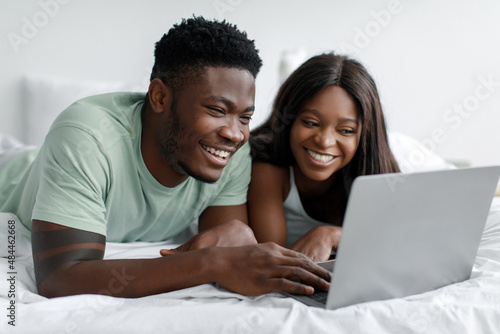 Laughing millennial black guy and woman looking at computer lie on white bed in bedroom