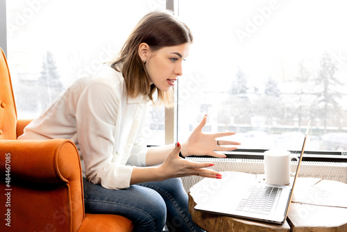 Irritated business woman having online conference with coworkers, discussing work troubles. Serious female entrepreneur is sitting on chair, using laptop, talking with colleagues at online meeting