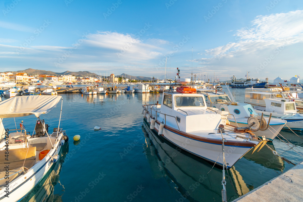 Colorful fishing boats line the harbor of the Greek island of Aegina, Greece at dusk, with the waterfront promenade and village in view.
