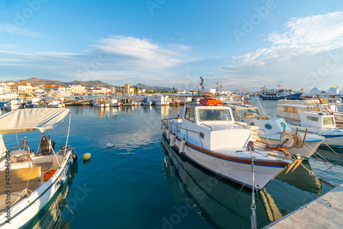 Colorful fishing boats line the harbor of the Greek island of Aegina, Greece at dusk, with the waterfront promenade and village in view. 