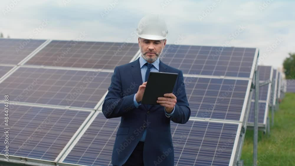 Successful Caucasian male businessman in suit and helmet stands in field with solar panels and focuses on typing using tablet. Successful Caucasian male businessman in suit and helmet stands in field with solar panels and focuses on typing using tablet.