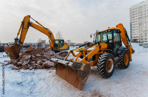 an excavator digs the ground at a construction site in winter against the background of a new house