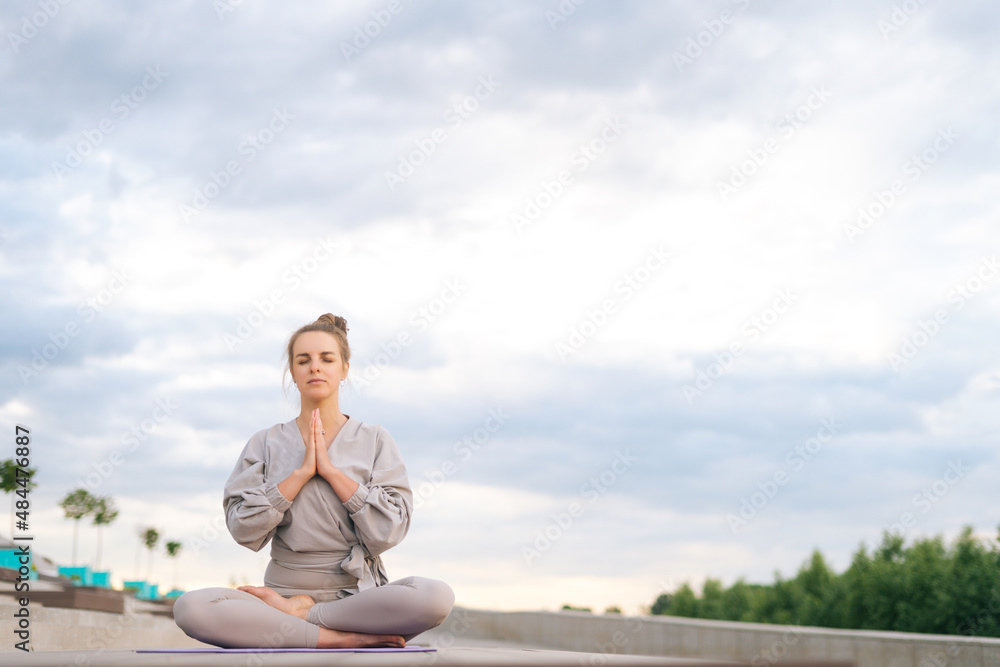 Portrait of calm Caucasian young woman practicing yoga performing namaste pose with closed eyes outside in city park. Pretty serene female sitting lotus position on yoga mat outdoors alone.