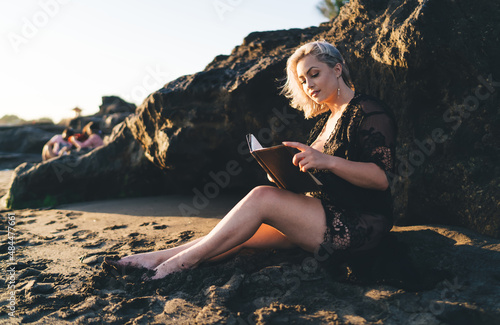 Beautiful female reading literature best seller sitting at sand and resting with hobby, charming Caucaisan woman in stylish wear holding education notepad analyzing book plot during vacations #484477661