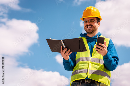 young caucasian male construction engineer smiling with a phone and notepad in hands.