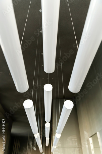 Modern design of lamps in the interior on the false ceiling of the hall. Volumetric composition of long cylindrical white fluorescent lamps.