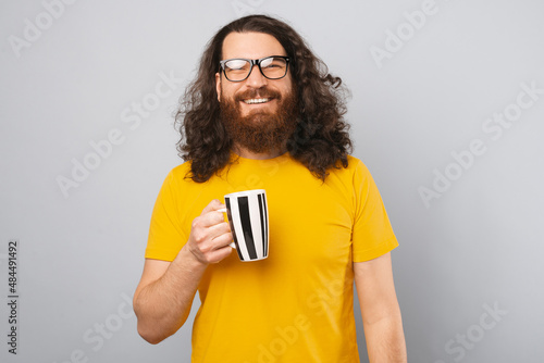 Curly bearded man is holding a mug while smiling at the camera. Portrait over grey background.