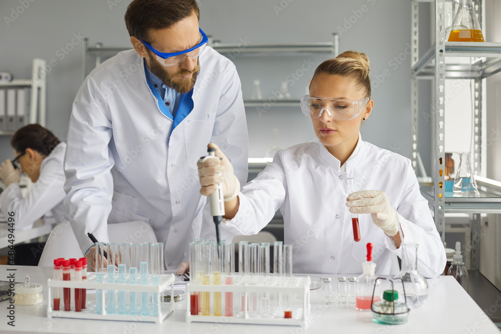 Two scientists working together on the analysis of blood samples while working in the laboratory. Male scientist observes a colleague who uses a pipette to add liquid to one of several test tubes. Two scientists working together on the analysis of blood samples while working in the laboratory. Male scientist observes a colleague who uses a pipette to add liquid to one of several test tubes.