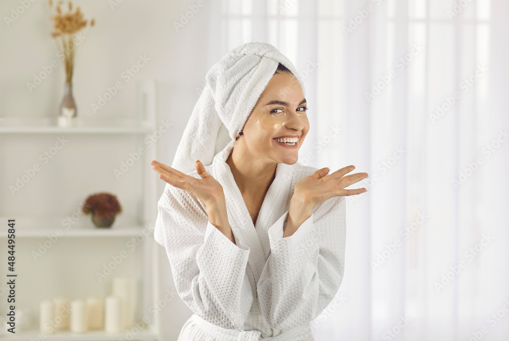 Portrait of happy woman wearing bathrobe and towel on her head performing her daily skin care routine at home. Woman with patches under her eyes spreads her arms and smiles broadly looking at camera. Portrait of happy woman wearing bathrobe and towel on her head performing her daily skin care routine at home. Woman with patches under her eyes spreads her arms and smiles broadly looking at camera.
