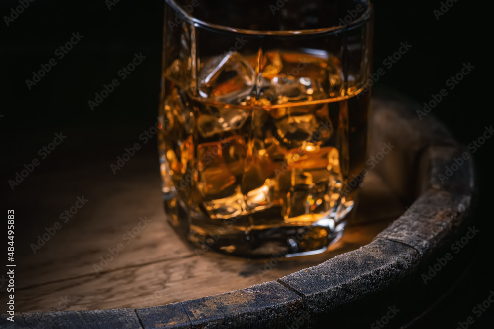 Glass of whiskey with ice blurry photo, selective focus on part of wooden barrel. Soft focus of photo of glass of brandy with ice close-up. Glass of whiskey with ice blurry photo, selective focus on part of wooden barrel. Soft focus of photo of glass of brandy with ice close-up.