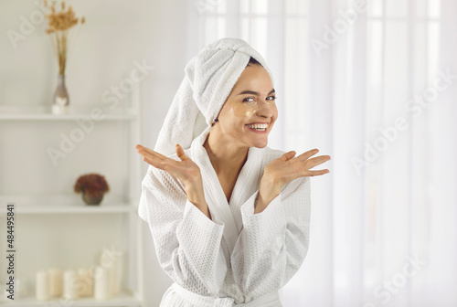 Portrait of happy woman wearing bathrobe and towel on her head performing her daily skin care routine at home. Woman with patches under her eyes spreads her arms and smiles broadly looking at camera.