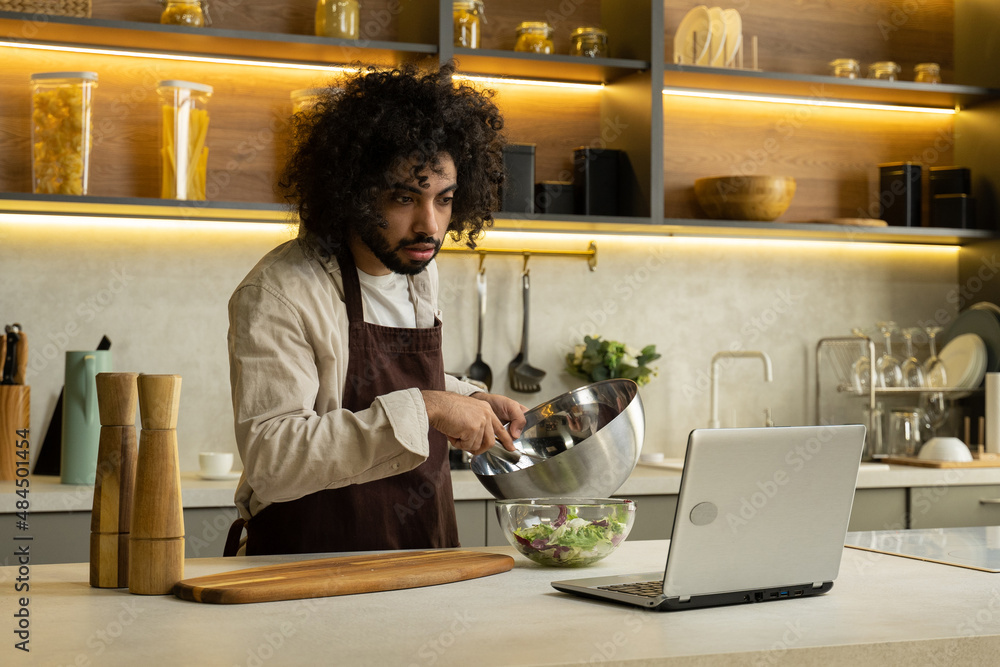 Egyptian man in apron with dark kinky hair cooks salad with lettuce in bowl watching recipe in laptop at table in modern kitchen Egyptian man in apron with dark kinky hair cooks salad with lettuce in bowl watching recipe in laptop at table in modern kitchen
