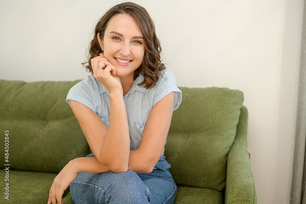 Healthy woman with brunette hair with glowing eyes, sitting on the pure comfortable sofa at modern apartment, dressed in casual clothes, put chin on the hand Healthy woman with brunette hair with glowing eyes, sitting on the pure comfortable sofa at modern apartment, dressed in casual clothes, put chin on the hand