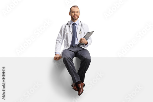 Male doctor smiling and sitting on a blank panel with a clipboard in his hands © Ljupco Smokovski