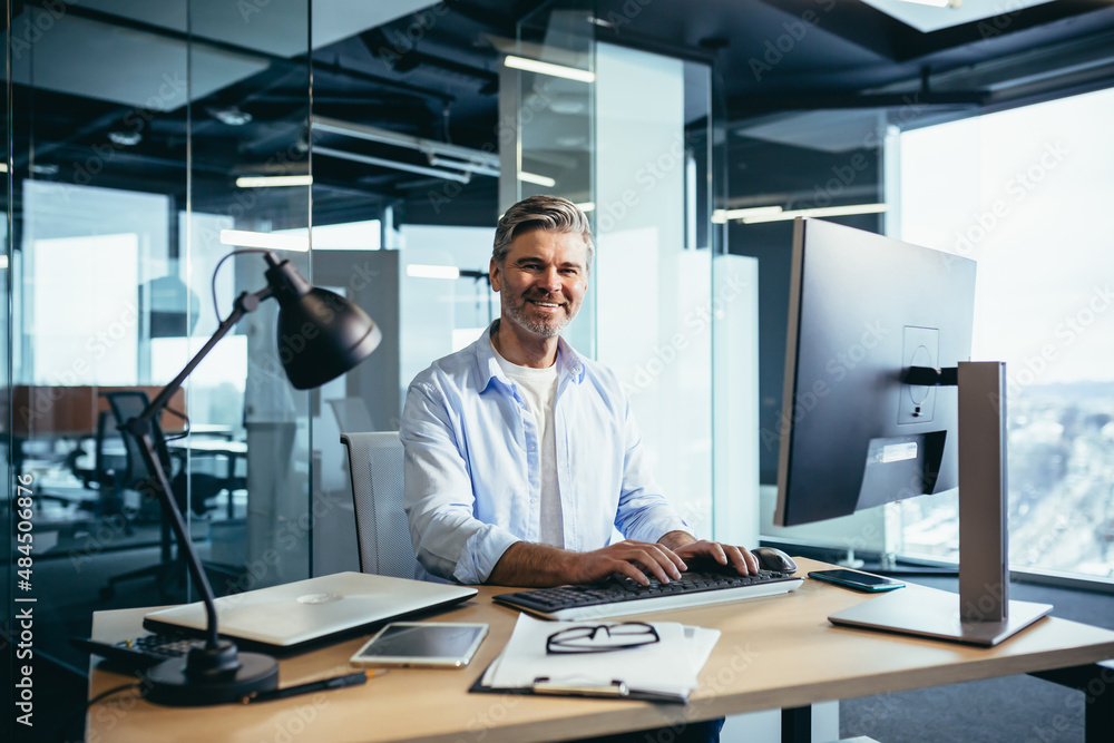 Portrait of a successful businessman, man rejoices and smiles, works at the computer, gray-haired and experienced looks at the camera Portrait of a successful businessman, man rejoices and smiles, works at the computer, gray-haired and experienced looks at the camera