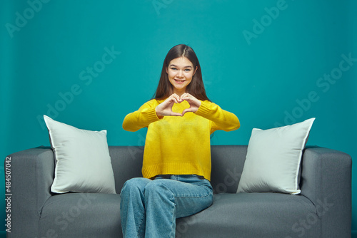 Friendly young girl show heart gesture, love sign, sitting on sofa. Blogger ask followers to put like. Charity, donation