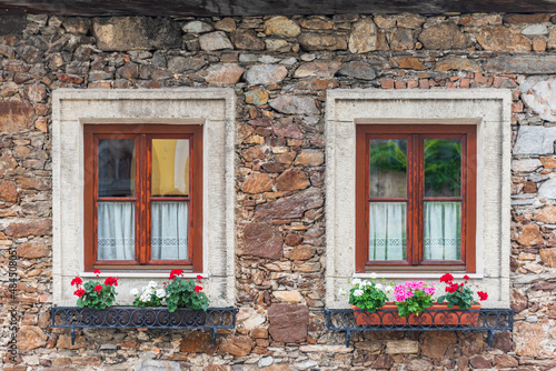 Fototapeta Naklejka Na Ścianę i Meble -  A wooden, brown window and a fragment of a stone wall of the building. Curtains hanging in the window, colorful flowers on the windowsill.