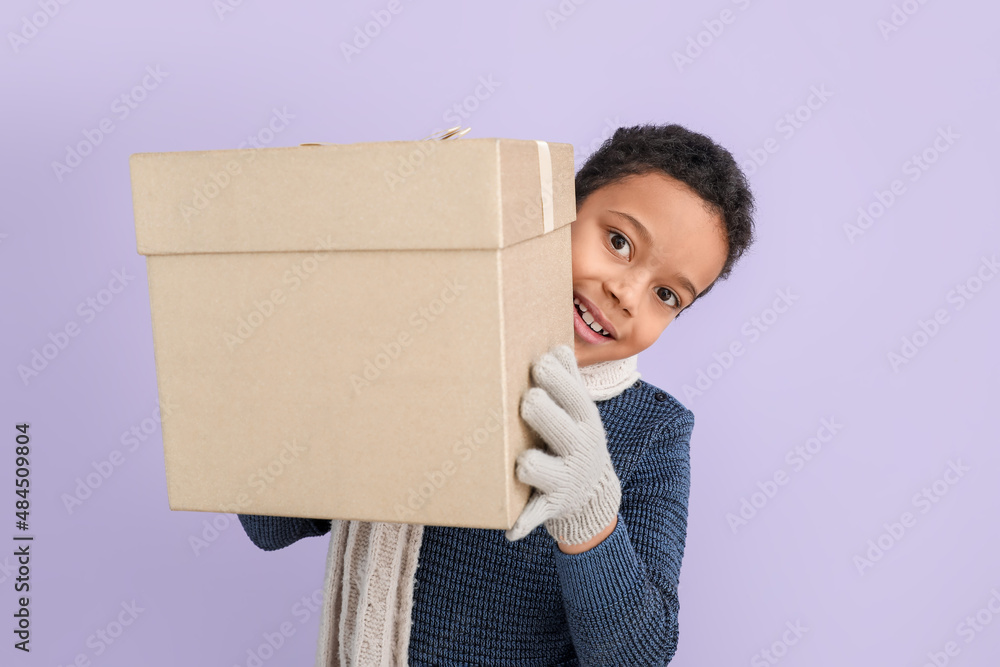 Cute little African-American boy in winter clothes and with Christmas gift on color background