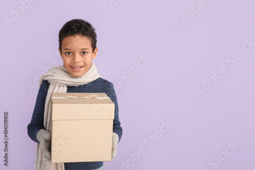 Cute little African-American boy in winter clothes and with Christmas gift on color background