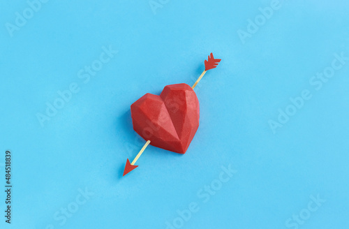 Red polygonal heart pierced by arrow on blue background. Concept of falling in love or Valentine's day. Copy space