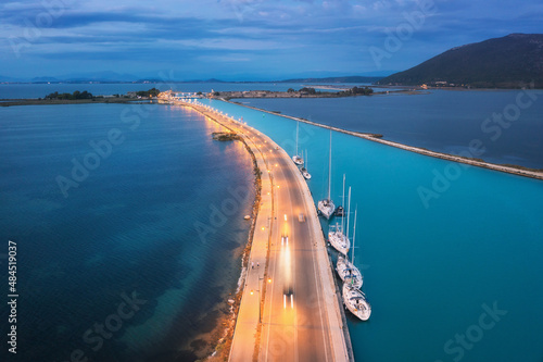 Aerial view of beautiful road near sea canal at night in summer in Lefkada, Greece. Top view of road, blurred cars, boat and yachts, city lights, azure water, mountain and cloudy sky at dusk. Travel 