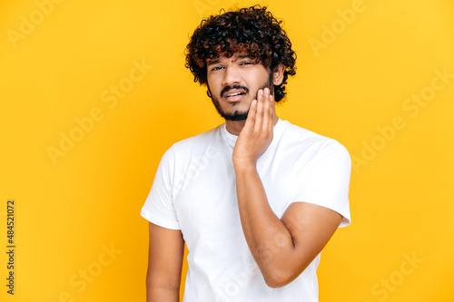 Toothache concept. Sad unhealthy indian or arabian guy touching cheek with hand with painful expression because of toothache or dental illness on teeth, standing on orange isolated background