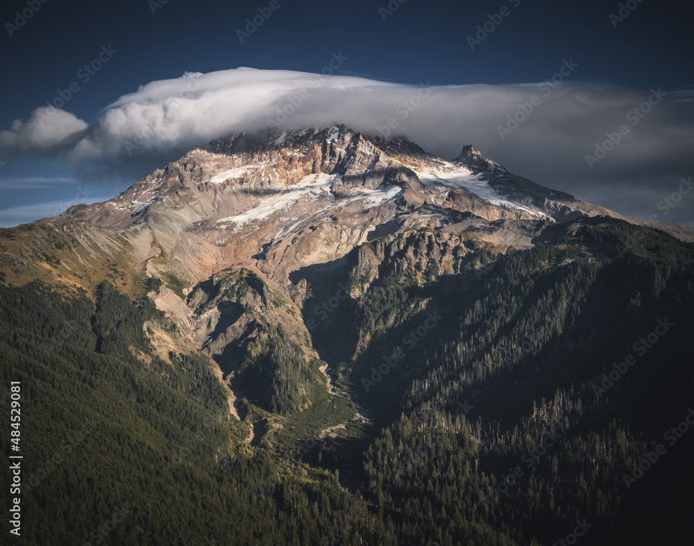A lenticular cloud hugs the top of snow covered Mt Hoot on a beautiful sunset in autumn. With a mostly blue sky there's a mountain range of craggy peaks below Hood and a line of evergreen trees