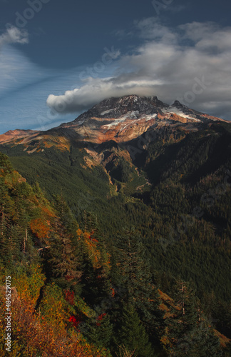 A lenticular cloud hugs the top of snow covered Mt Hoot on a beautiful sunset in autumn. With a mostly blue sky there's a mountain range of craggy peaks below Hood and a line of evergreen trees