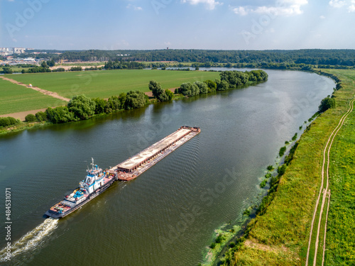 Fotografija Towboat pushes barge with sand along calm river past fields