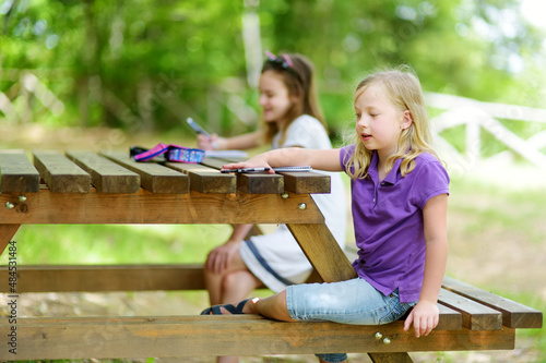 Two happy sisters drawing together at the wooden table outdoors. Little girls sketching outside on beautiful summer day.