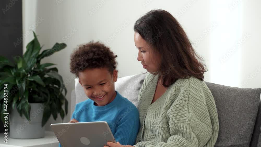 Glad and positive woman asian mom sitting on comfort couch inside bright light living modern room with her glad african mixed races son. They looking at tablet together, making wide beaming smile. 4k Glad and positive woman asian mom sitting on comfort couch inside bright light living modern room with her glad african mixed races son. They looking at tablet together, making wide beaming smile. 4k