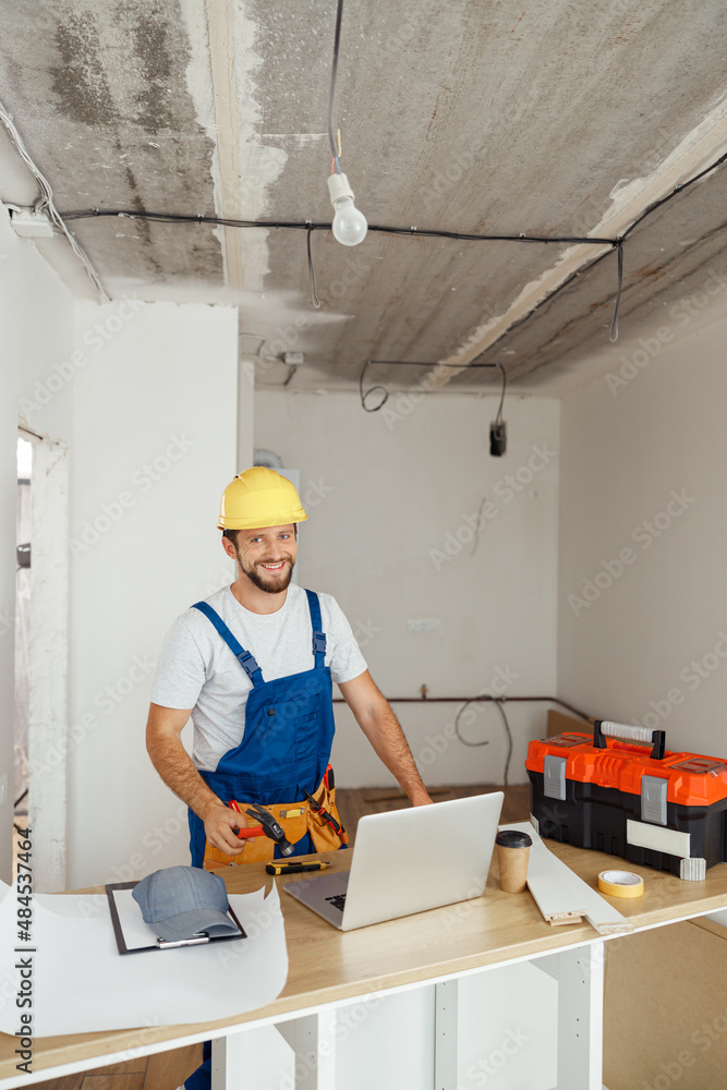 Happy repairman worker in uniform wearing tool belt and hardhat holding hammer and smiling at camera while standing indoors Happy repairman worker in uniform wearing tool belt and hardhat holding hammer and smiling at camera while standing indoors