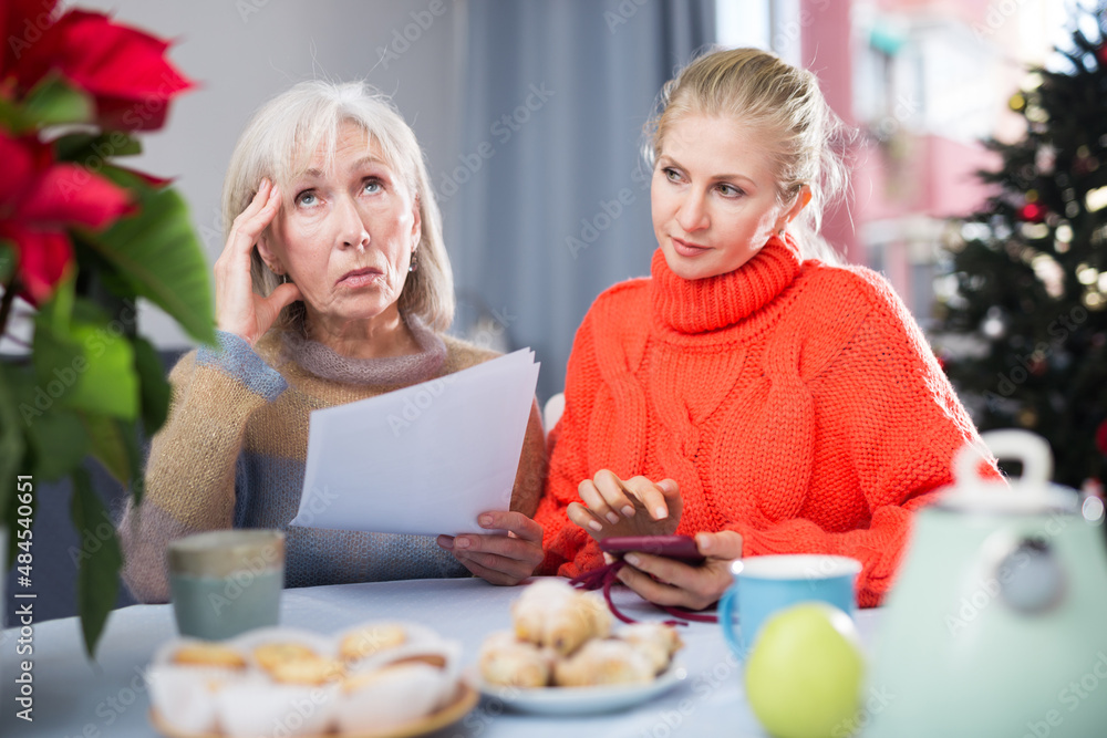 Mature woman and her adult daughter, who came to visit her before Christmas, study an important document sitting at a table, ..dialing a number on a mobile phone that is listed there