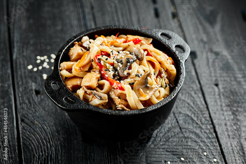 Stir fried udon noodles in wok. Udon noodle with chicken and mushrooms on wooden background. Asian noodles on black table with ingredients. Wok menu for japanese restaurant.