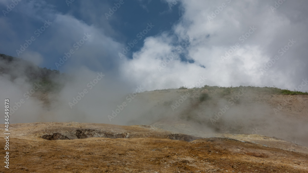 The soil in the geothermal valley is covered with orange sulfur deposits. Steam and smoke from fumaroles rise into the blue sky. The slopes of the mountains are hidden in a haze. Kamchatka