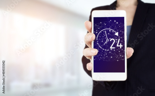 Technical Support Center Customer Service Business Concept. Hand hold white smartphone with digital hologram 24 7 all day all night sign on light blurred background.