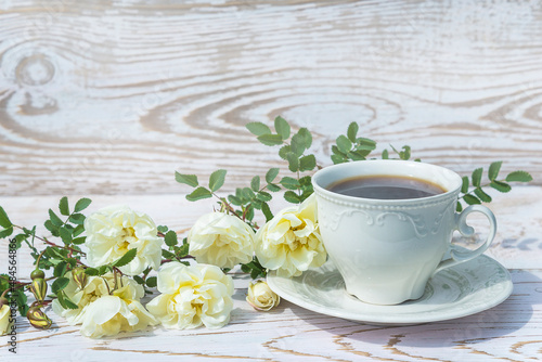 White cup of coffee and twig with roses flowers on white rustic wooden background