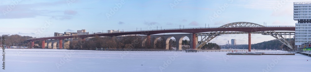 Naklejka premium Panorama view of two train bridges, old and new, between the districts Årstadal and Södermalm, a snow covered and frozen lake Årstaviken a cold sunny winter day in Stockholm
