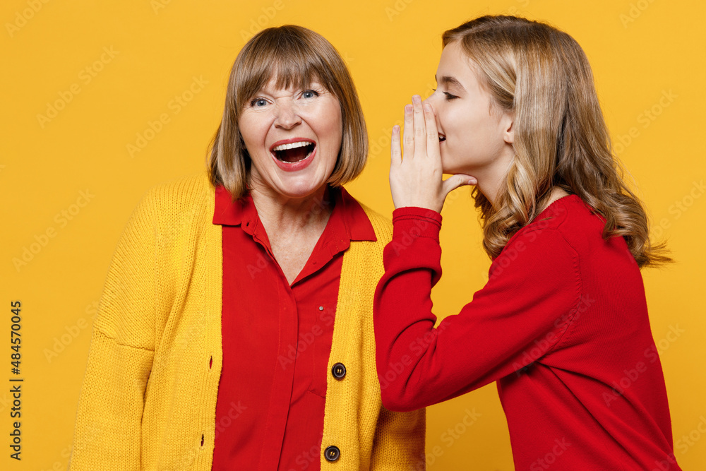 Happy woman 50s in red shirt have fun with teenager girl 12-13 years old. Grandmother granddaughter whispering gossip and tells secret behind her hand sharing news isolated on plain yellow background