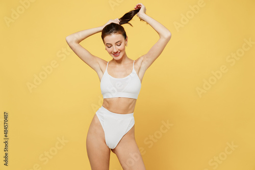 Smiling lovely pretty nice attractive young brunette woman 20s wearing white underwear with perfect fit body standing ties hair in ponytail isolated on plain yellow color background studio portrait