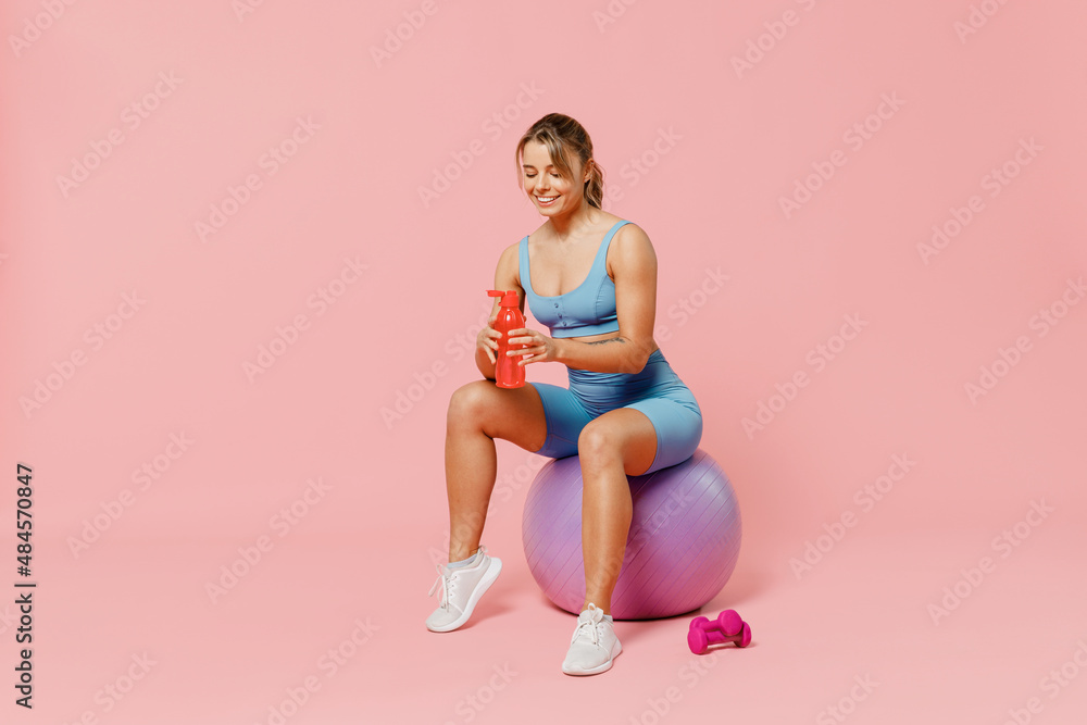 Full body young athletic fitness trainer instructor woman wear blue tracksuit spend time in home gym sit on fitball drink water isolated on pastel plain light pink background Workout sport concept. Full body young athletic fitness trainer instructor woman wear blue tracksuit spend time in home gym sit on fitball drink water isolated on pastel plain light pink background Workout sport concept.