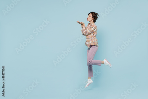 Full body side profile view fun young happy woman 20s wearing casual brown shirt jump high blow air kiss isolated on pastel plain light blue color background studio portrait. People lifestyle concept.