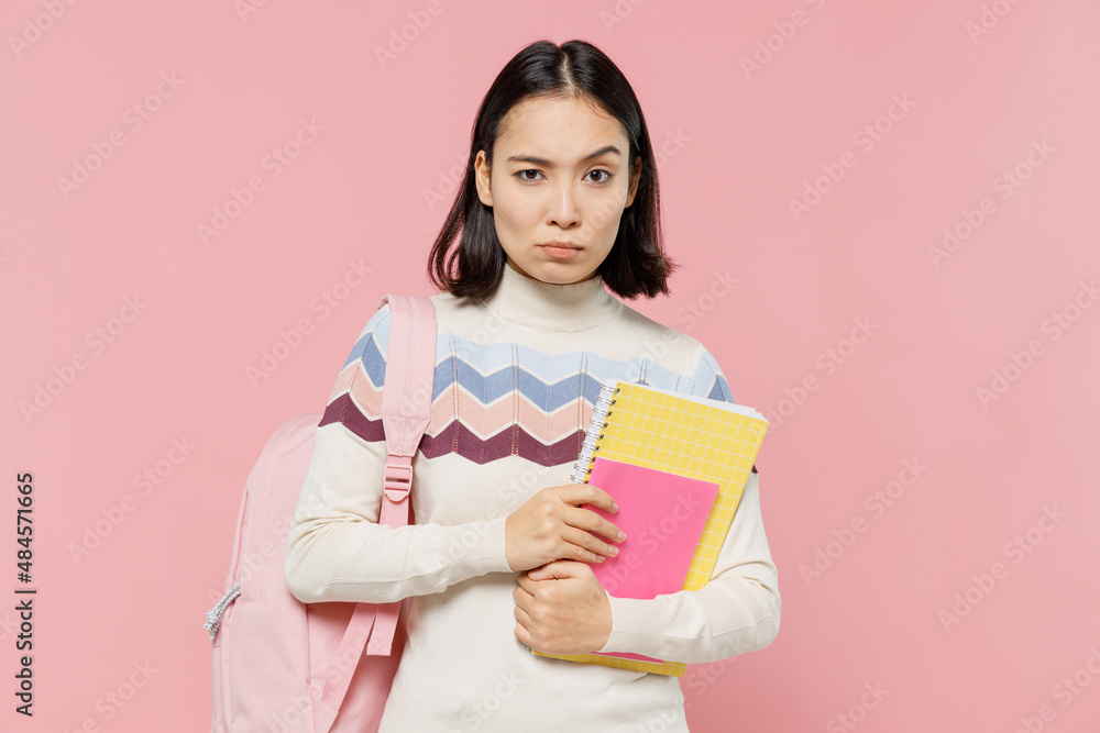Serious strict sad teen student girl of Asian ethnicity wearing sweater backpack hold books look camera isolated on pastel plain light pink color background Education in university college concept Serious strict sad teen student girl of Asian ethnicity wearing sweater backpack hold books look camera isolated on pastel plain light pink color background Education in university college concept
