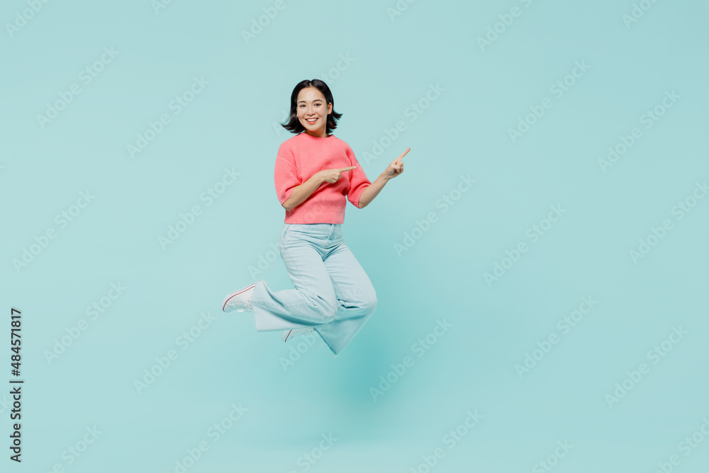 Full body young smiling happy woman of Asian ethnicity 20s wear pink sweater jump high point index finger camera on you isolated on pastel plain light blue background studio. People lifestyle concept.