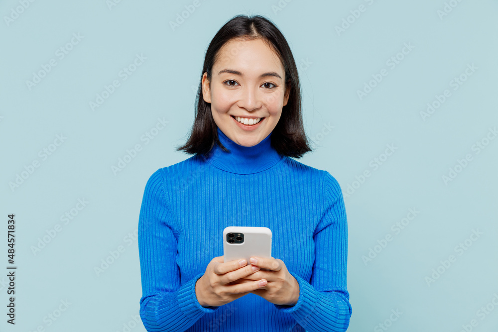 Smiling fancy young woman of Asian ethnicity 20s years old wears blue shirt hold in hand use mobile cell phone typing browsing chatting isolated on plain pastel light blue background studio portrait. Smiling fancy young woman of Asian ethnicity 20s years old wears blue shirt hold in hand use mobile cell phone typing browsing chatting isolated on plain pastel light blue background studio portrait.
