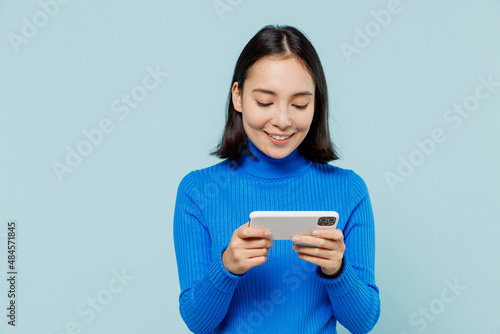 Young woman of Asian ethnicity 20s wears blue shirt using play racing app on mobile cell phone hold gadget smartphone for pc video games isolated on plain pastel light blue background studio portrait.